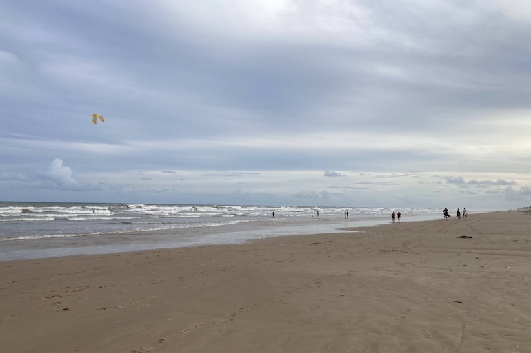 Praia de Aruanã, com suas areias escuras e um kite surf na água, no final da tarde. Poucas pessoas caminhando na areia.