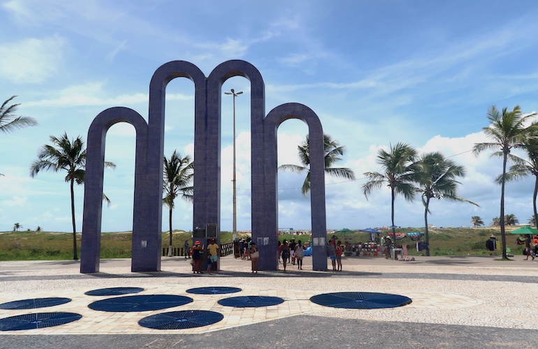 Monumento Arcos da Atalaia, no calçadão da orla, com a praia ao fundo.