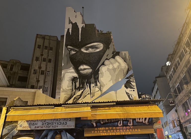 Prédio grafitado, com desenho de mulher vestindo máscara preta, demonstrando a vocação artística e cultural do bairro Psyri, uma das melhores áreas de Atenas para mochileiros e viajantes econômicos.